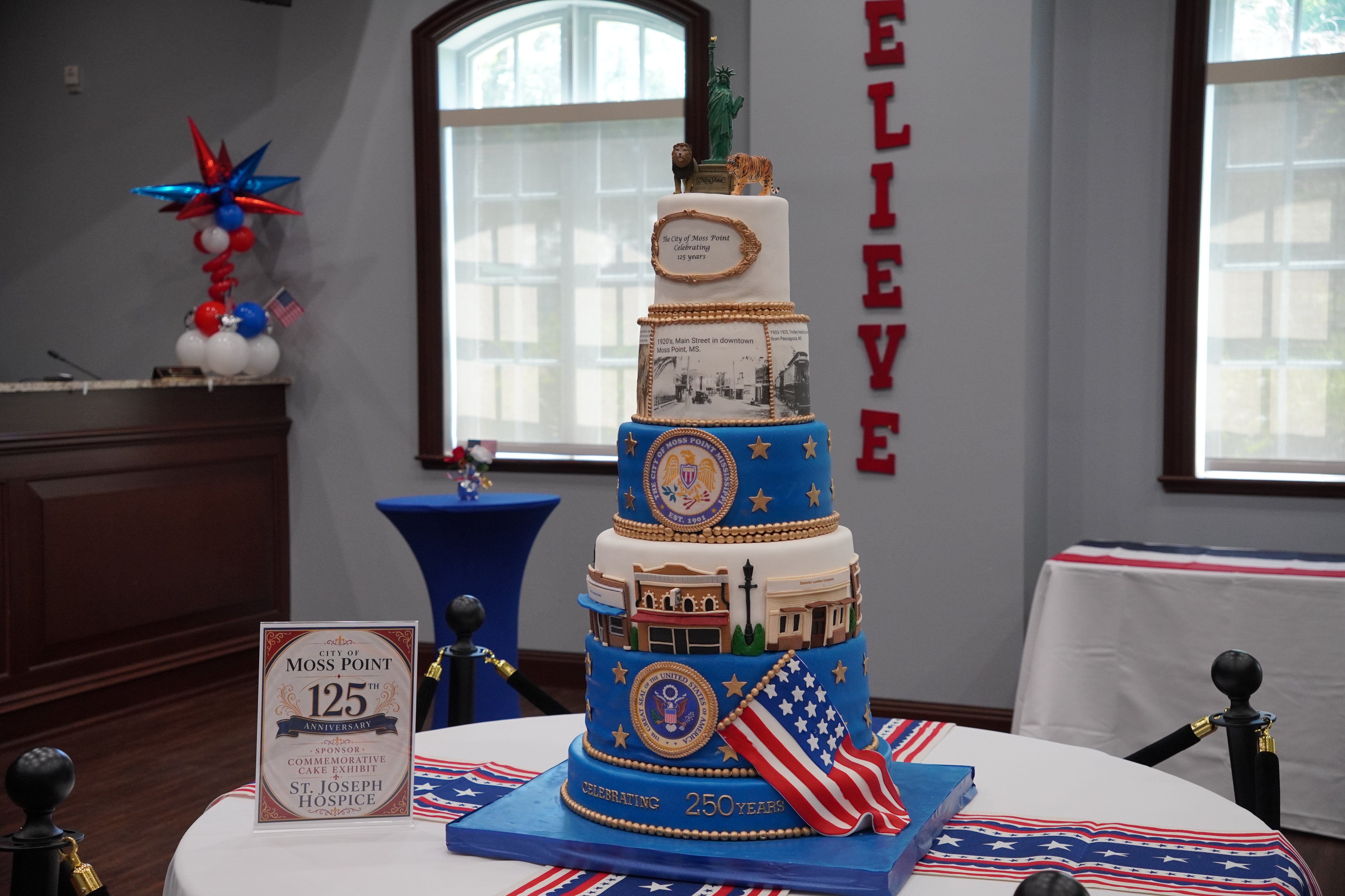 A multi-tier patriotic celebration cake honoring Moss Point's 125th anniversary and America's 250th Semiquincentennial, displayed at River Jamboree 2026 with the city's BELIEVE installation in the background